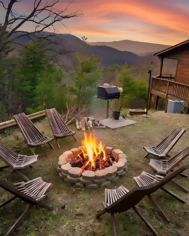 Fire pit glowing at sunset beside the cabin, with chairs, the charcoal grill and the mountain ridge behind
